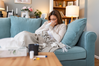 sick woman lying on couch blowing nose with tissue next to table with tea mug, thermometer, and medicine